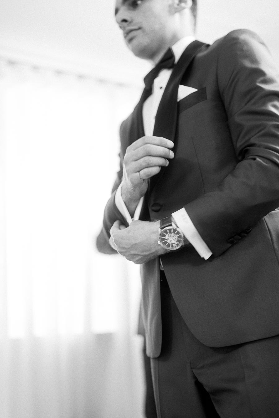 Groom in a black tuxedo adjusts his jacket, with cufflinks, watch, and bow tie visible in soft black and white.