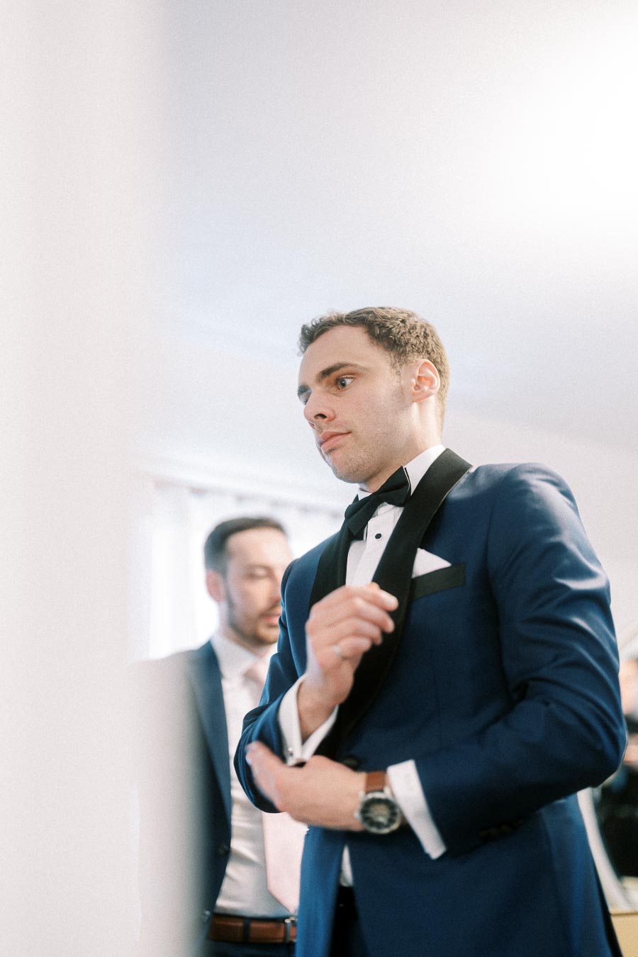 Groom in a navy tuxedo adjusts his cuff while getting ready indoors with a groomsman behind him.