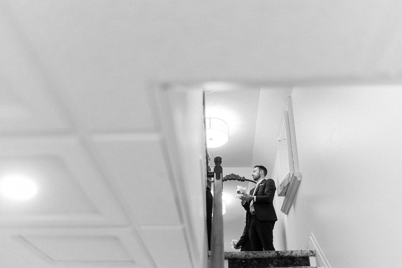 Black-and-white view of a suited wedding guest standing on a staircase holding flowers.