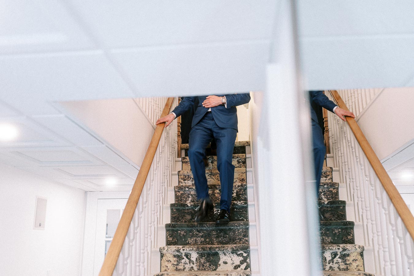 Groom in a blue suit descends a carpeted staircase, partly reflected in a wall mirror.