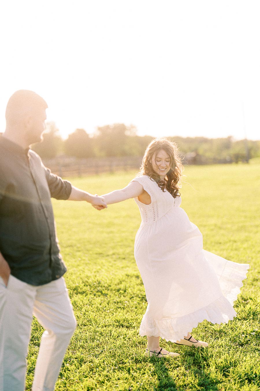 Expectant couple holding hands in a sunlit field at The Green Acres