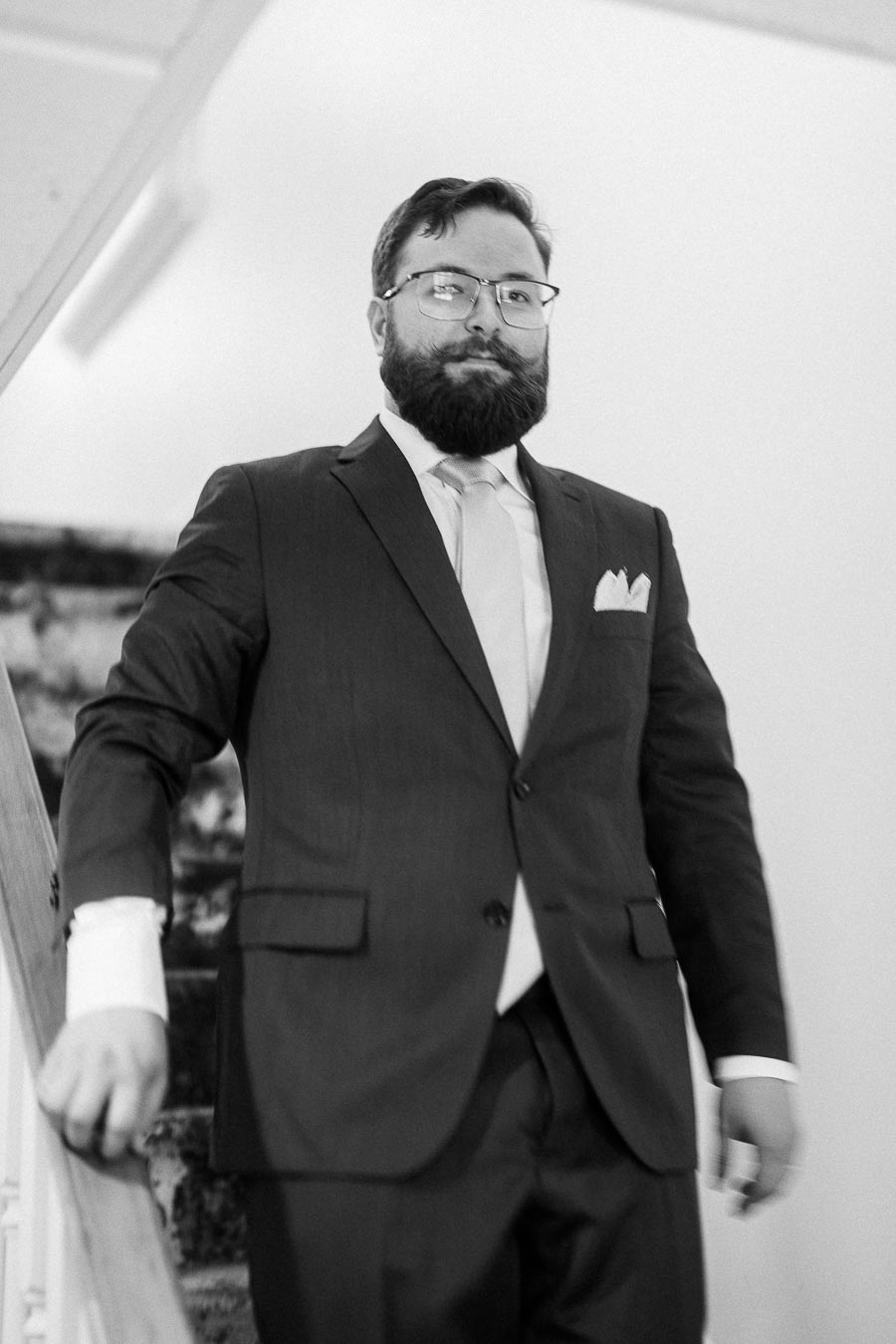 Black-and-white portrait of Rawle in a suit and tie, standing by a stair railing indoors.
