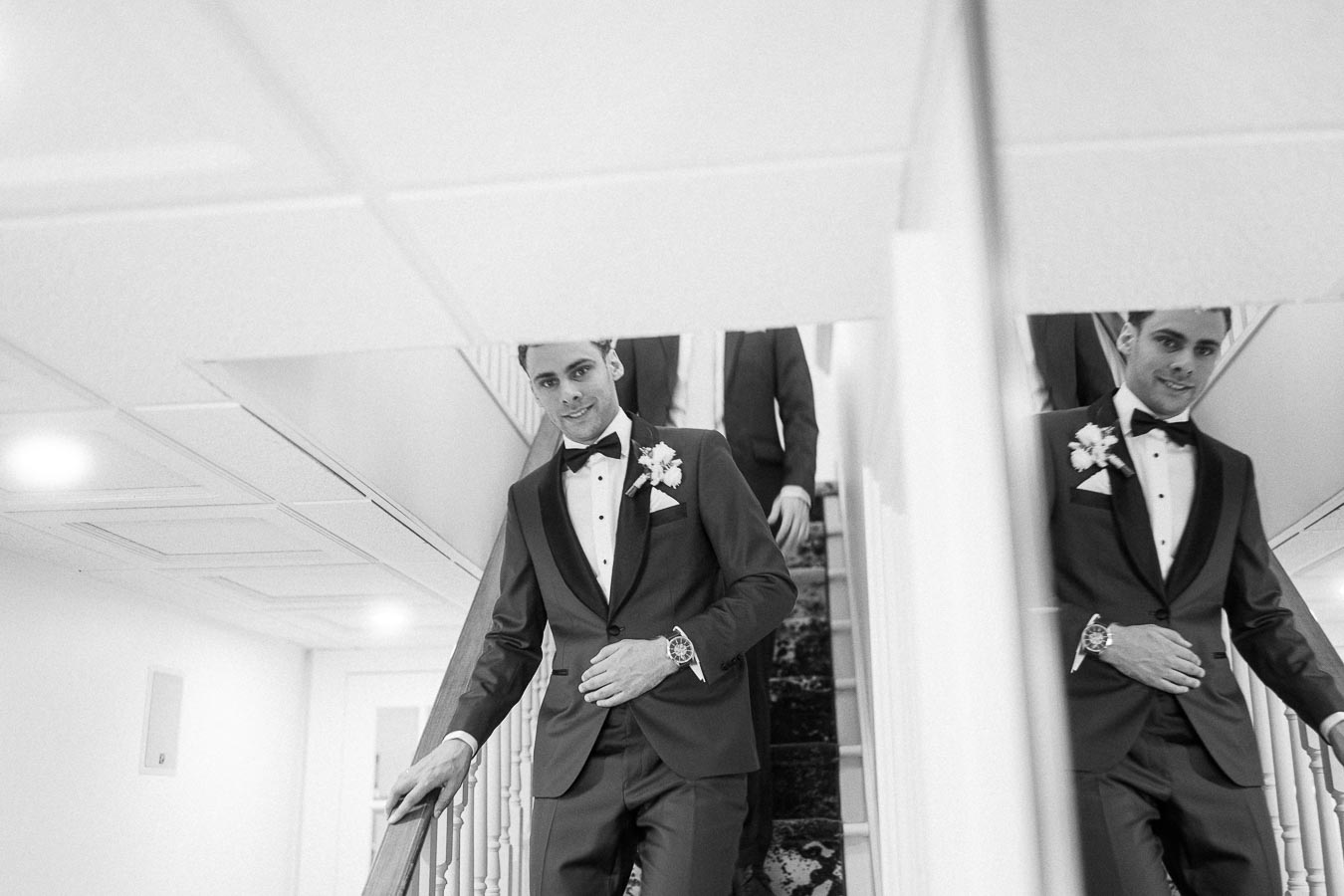 Groom in a tuxedo descends an indoor staircase, reflected in a wall mirror beside the railing.