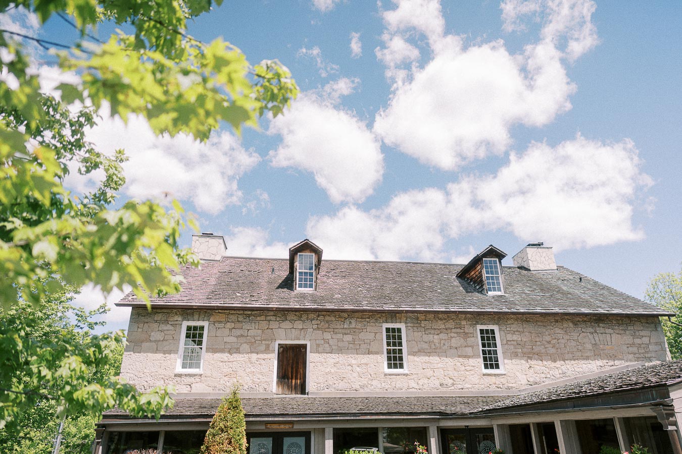 Stone building at Ancaster Mill under a blue sky with leafy green branches in the foreground.