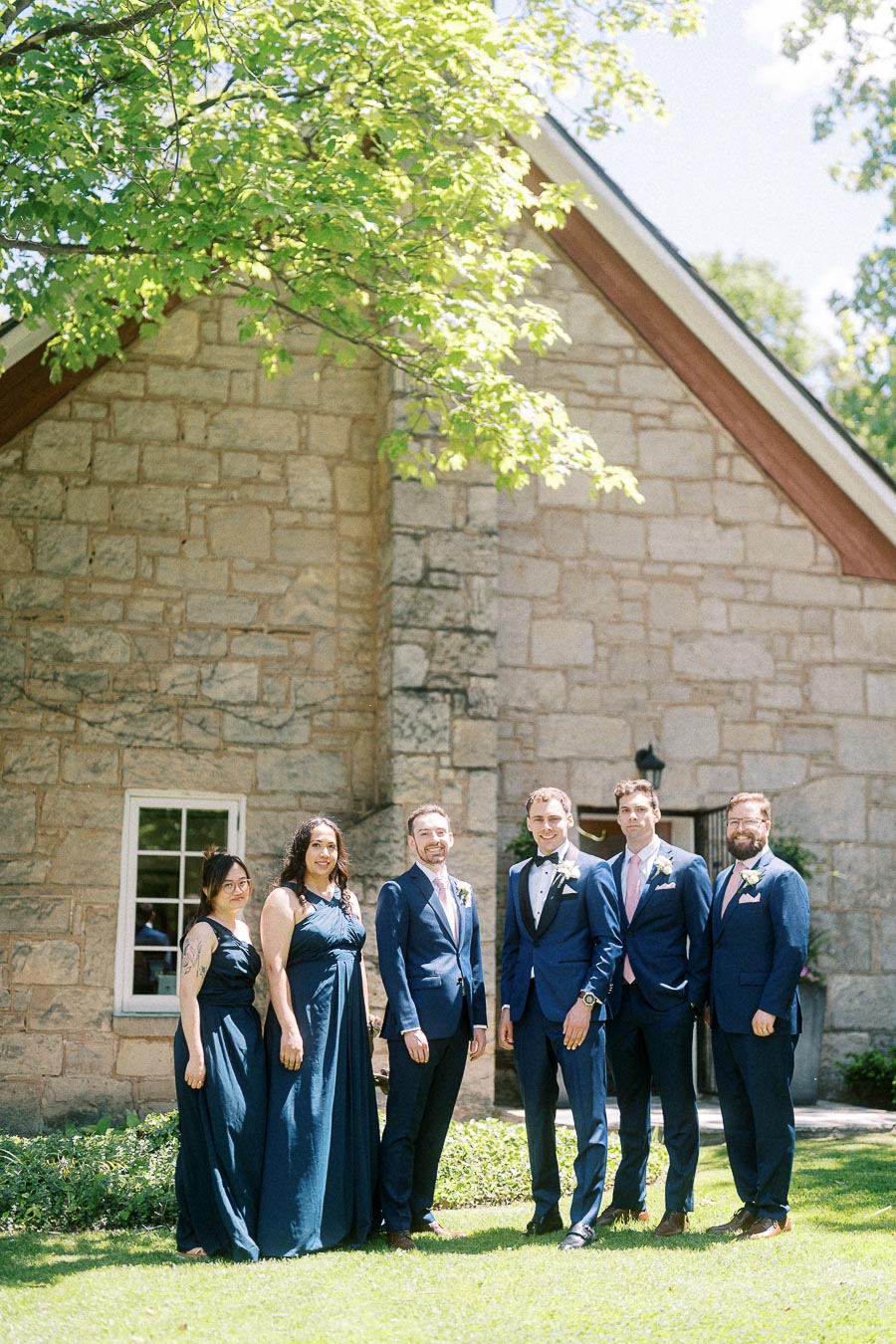 Wedding party in navy attire standing on the lawn beside a stone chapel at Ancaster Mill