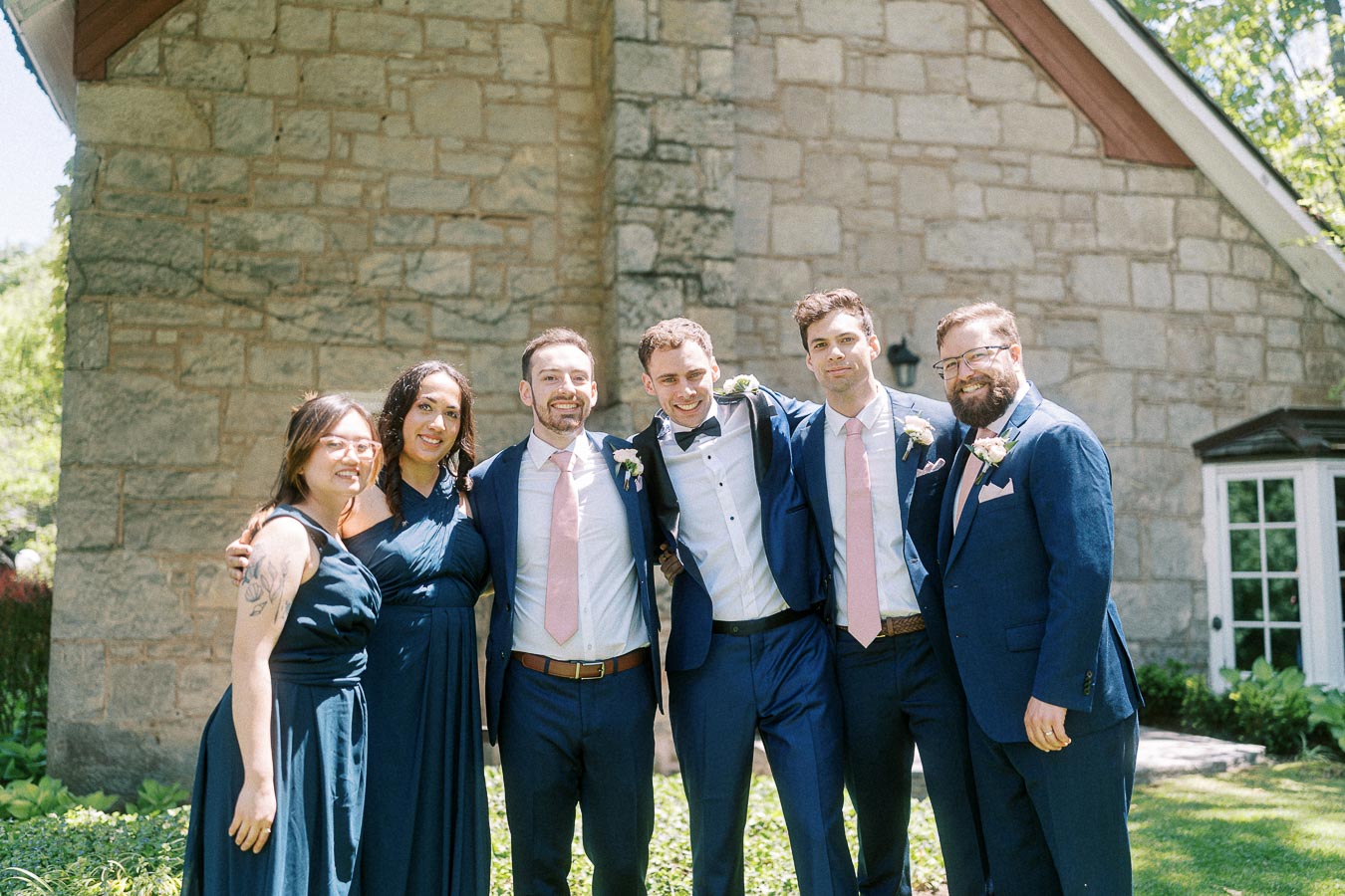 Six wedding party members in navy attire pose arm in arm outside a stone building at Ancaster Mill.