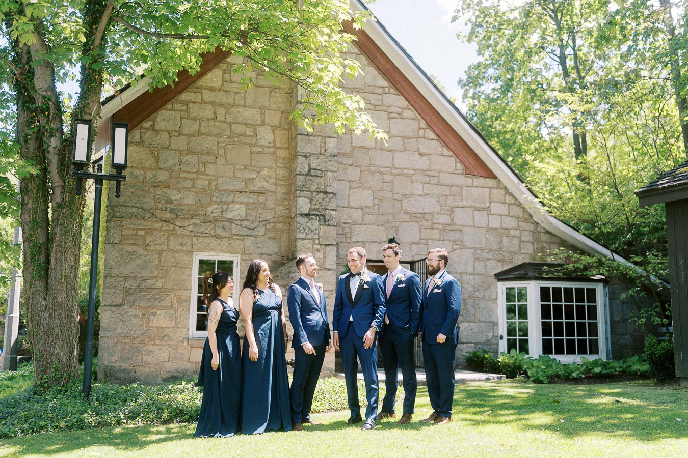 Wedding party in navy attire standing on grass beside a stone building and trees at Ancaster Mill.