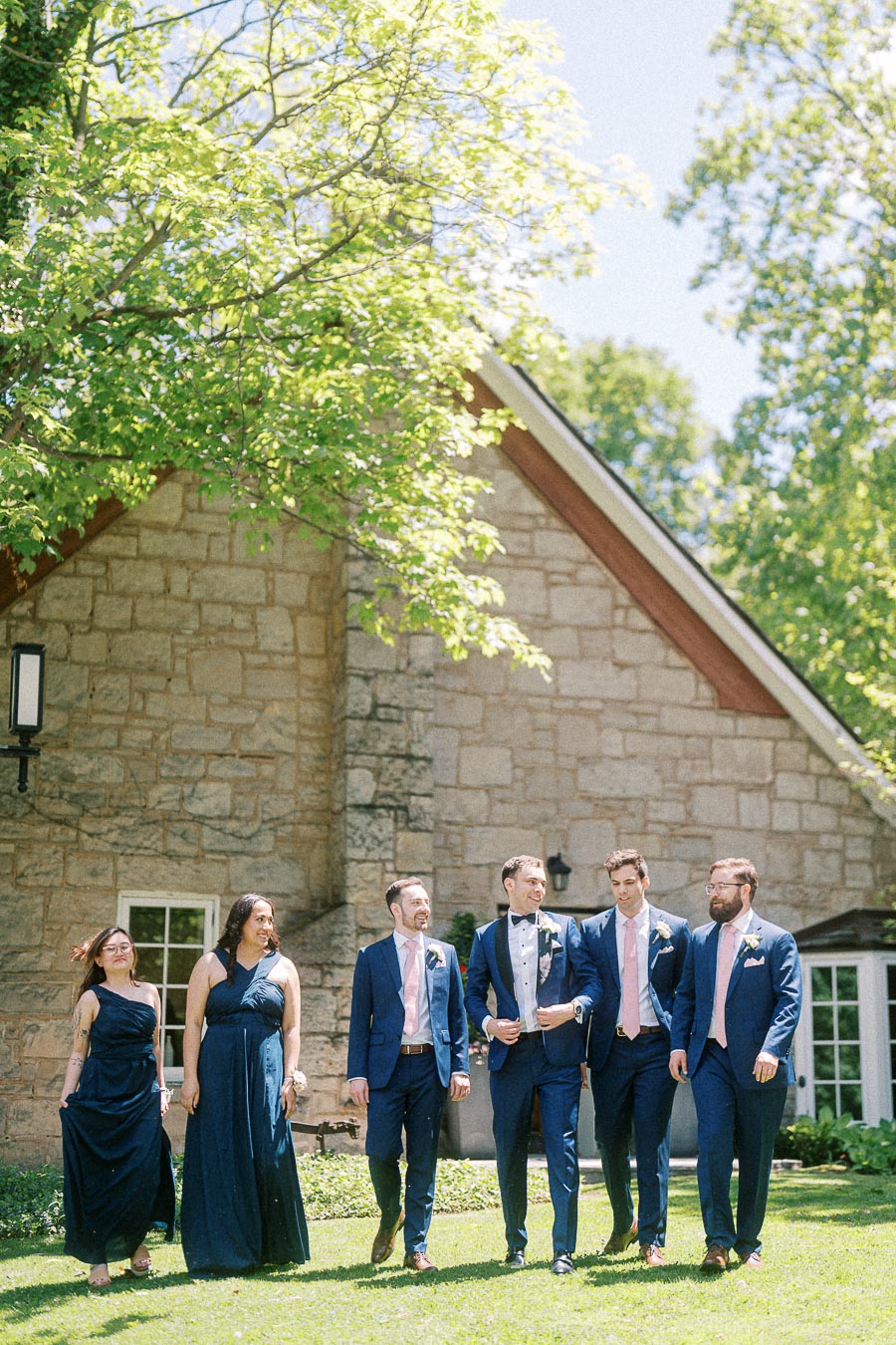 Wedding party in navy attire walking on the lawn beside a stone building at Ancaster Mill.