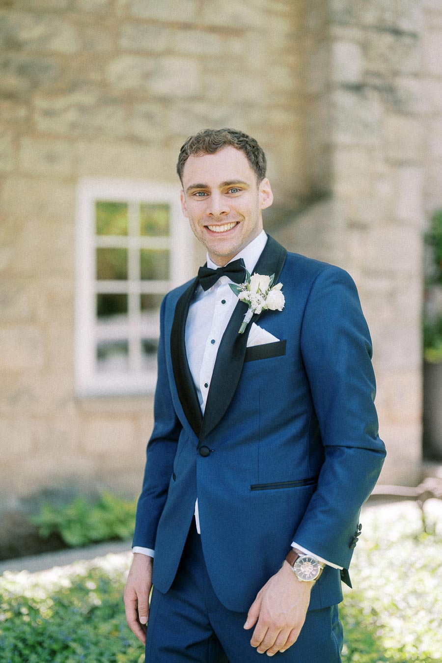 Groom in a blue tuxedo with black lapels smiles outdoors beside a stone building and window.