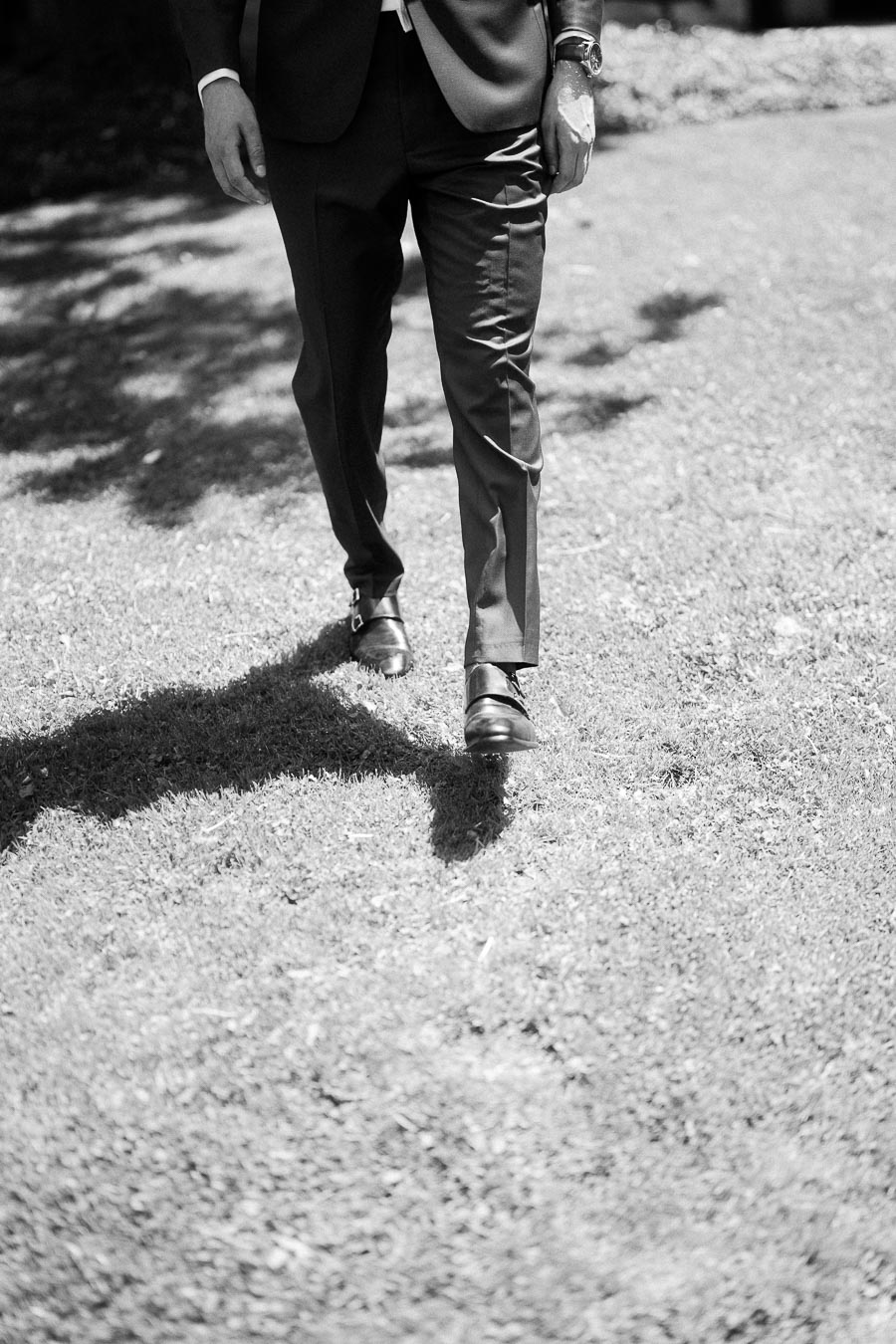 Lower half of a groom in a dark suit walking across sunlit grass in black-and-white