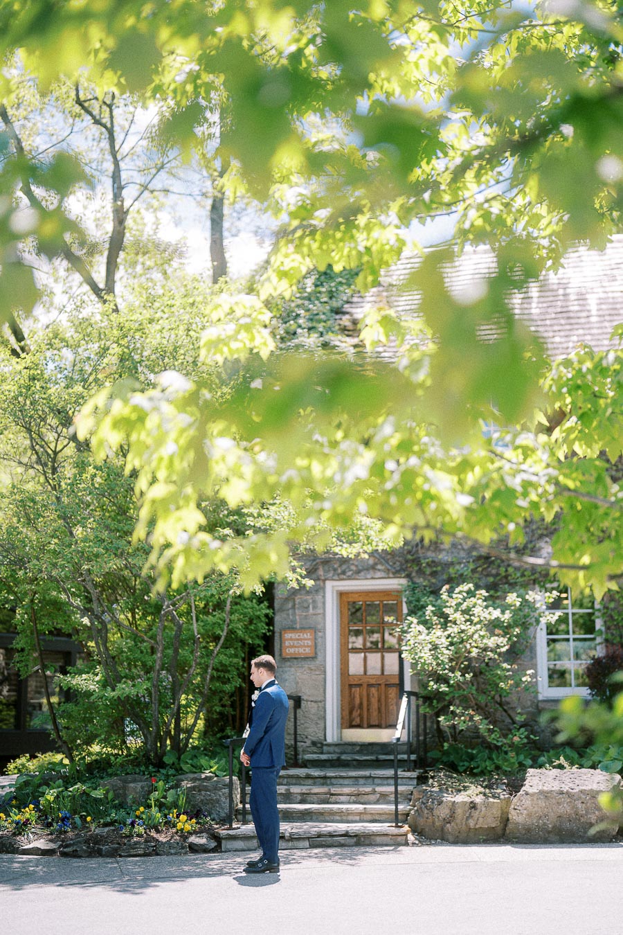 Groom in a blue suit stands outside a stone cottage entrance framed by green spring leaves.