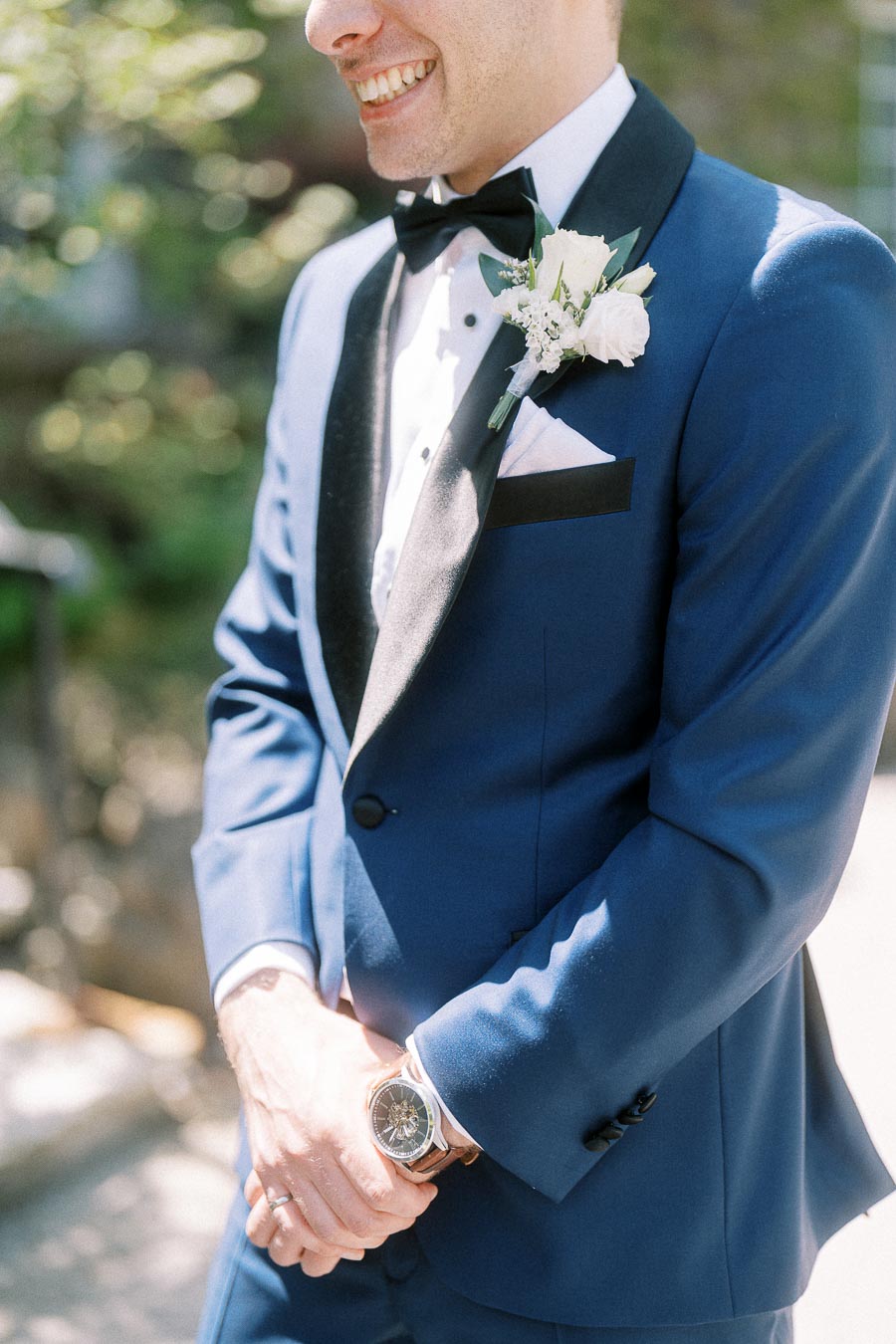 Groom in a navy tuxedo with black bow tie and white boutonniere standing outdoors in sunlight