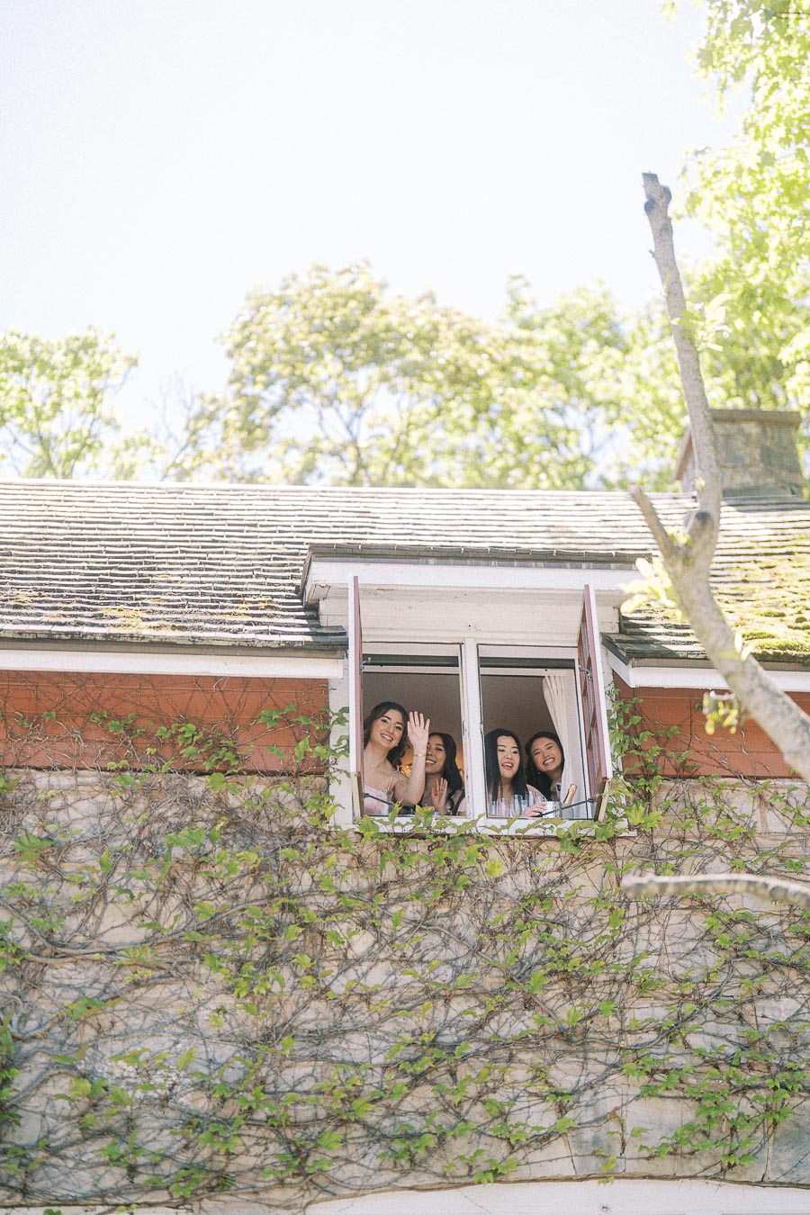 Four women wave from an upstairs window of a vine-covered stone building in bright spring light.
