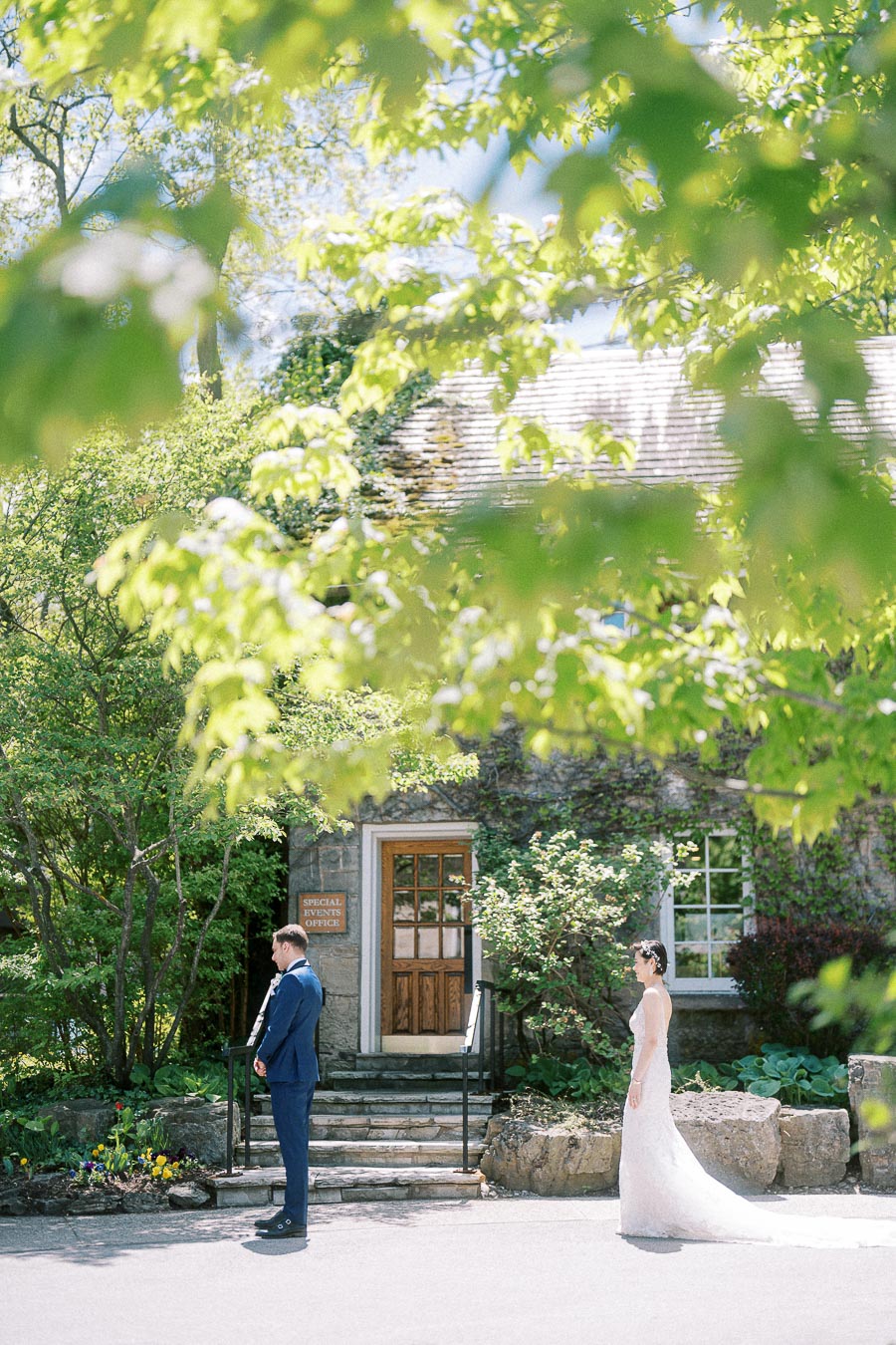 Bride approaches groom from behind for their first look outside a stone building framed by green leaves.