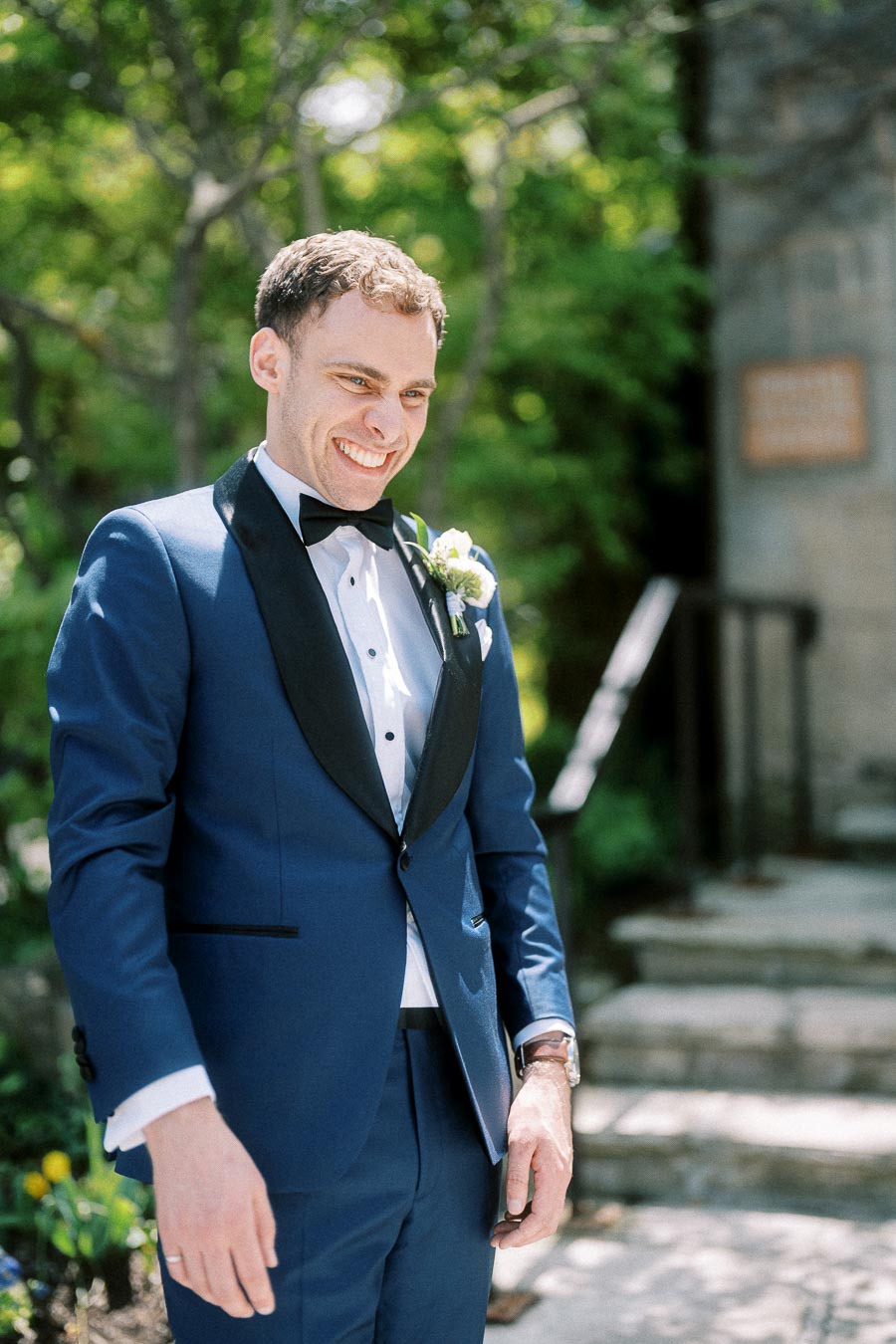 Rawle smiling in a blue tuxedo with boutonniere outdoors near stone steps and greenery.
