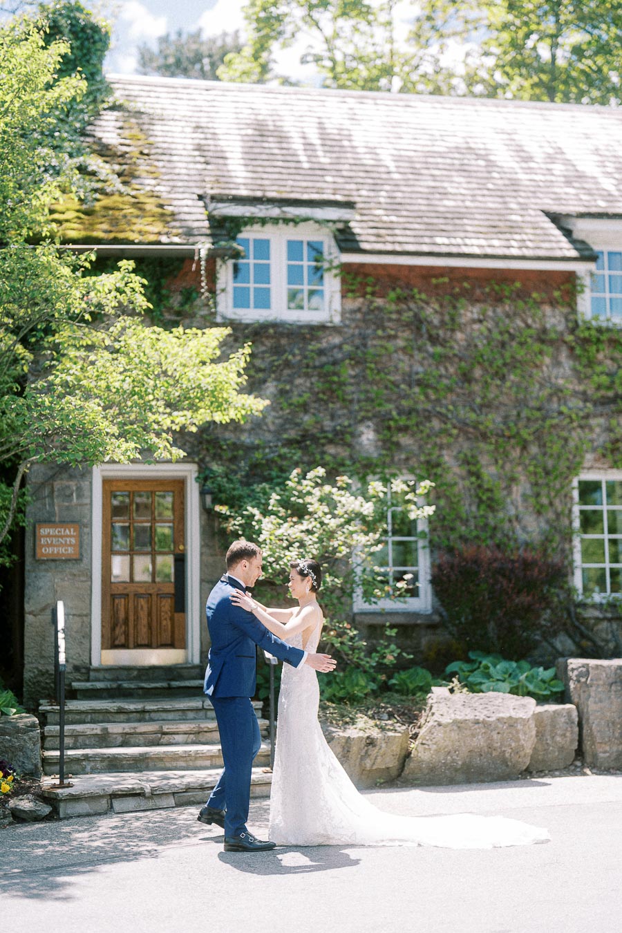 Bride and groom embrace outside the ivy-covered Ancaster Mill building on a sunny wedding day.