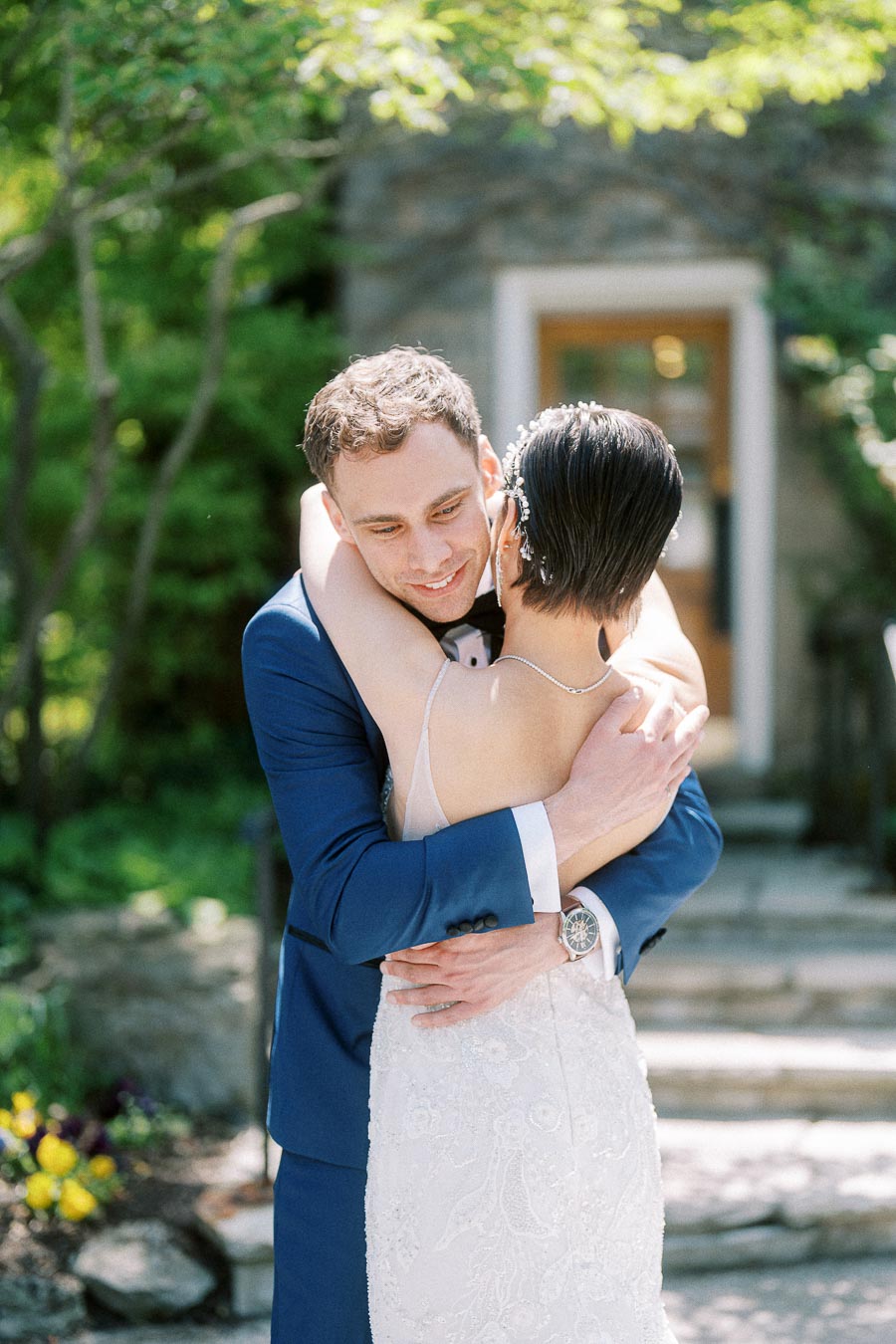 Rawle smiles while embracing Miffy in her beaded wedding dress outdoors among greenery and stone steps.