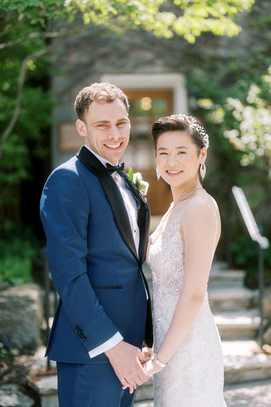 Miffy and Rawle smiling and holding hands outdoors in wedding attire beside greenery and stone steps.