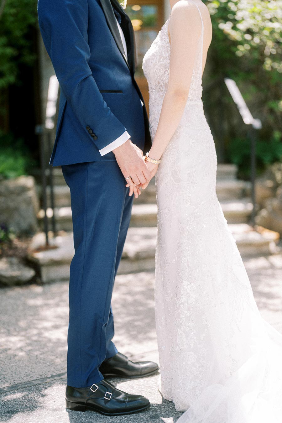 Bride and groom holding hands outdoors, showing a blue suit and lace wedding dress in soft sunlight.