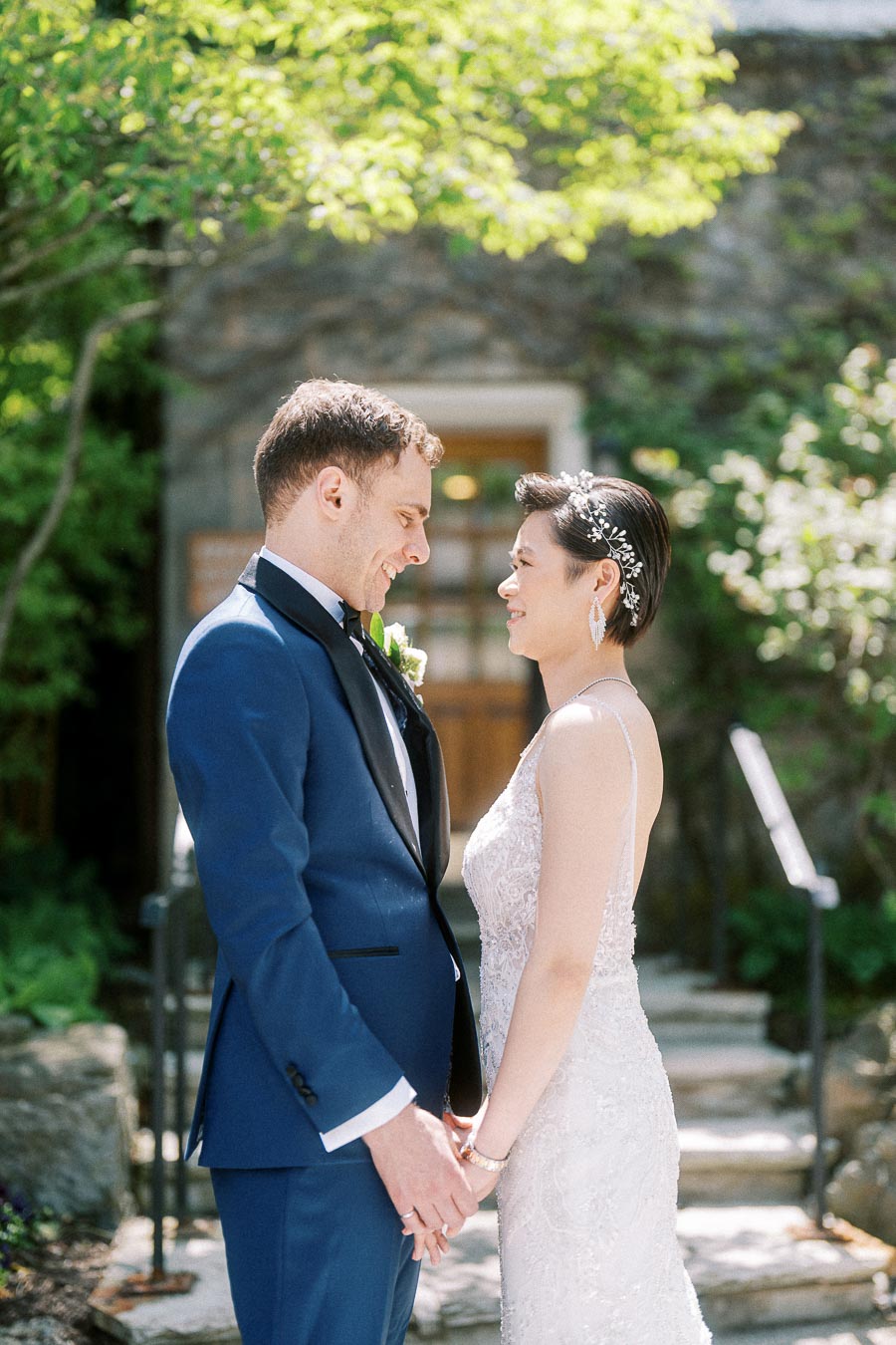 Miffy and Rawle smiling and holding hands outdoors in wedding attire near a stone building and greenery.