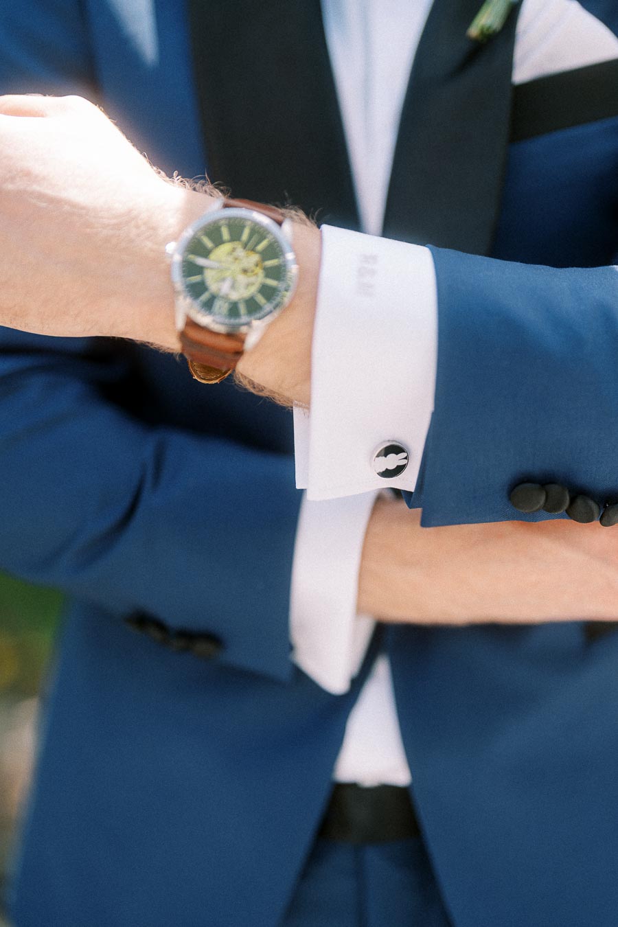 Close-up of a groom's blue suit sleeve, wristwatch, white cuff, and black cufflink outdoors