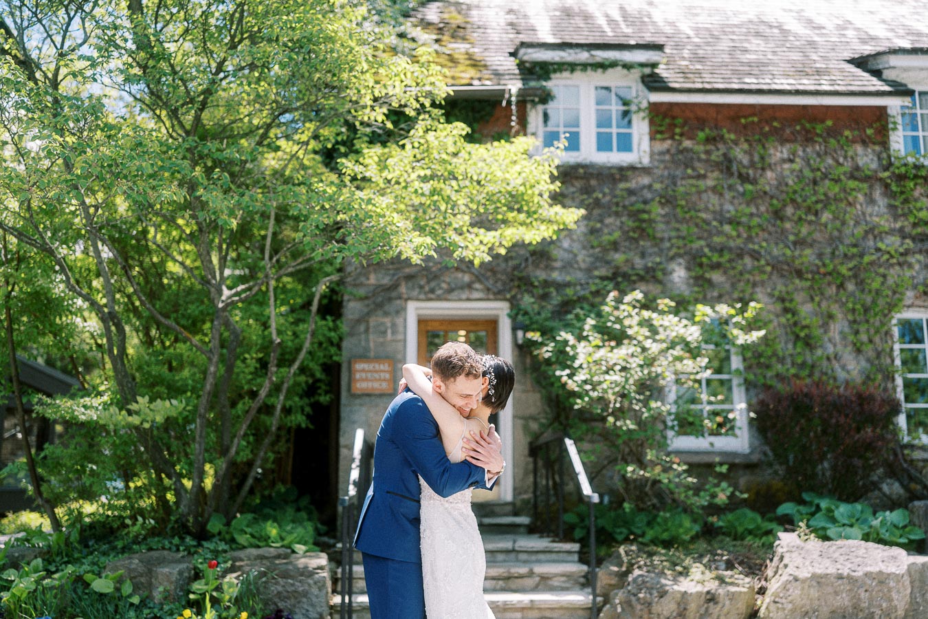 Miffy and Rawle embrace outside a stone, vine-covered building surrounded by trees at Ancaster Mill.