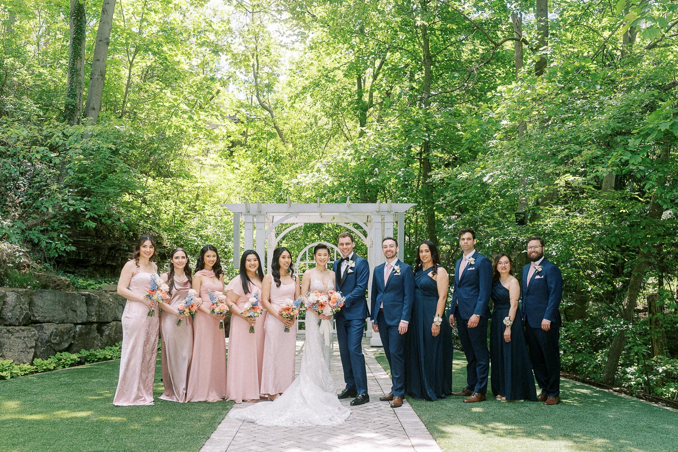 Miffy, Rawle, and their wedding party stand beneath a white garden arbor surrounded by green trees.