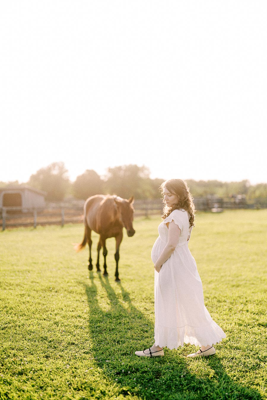Pregnant woman in a white dress standing in a pasture with a horse