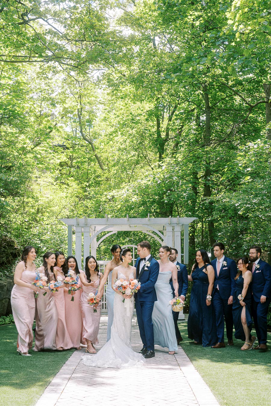 Wedding party in blush and navy stands with the bride and groom beneath a white arbor in a leafy garden.