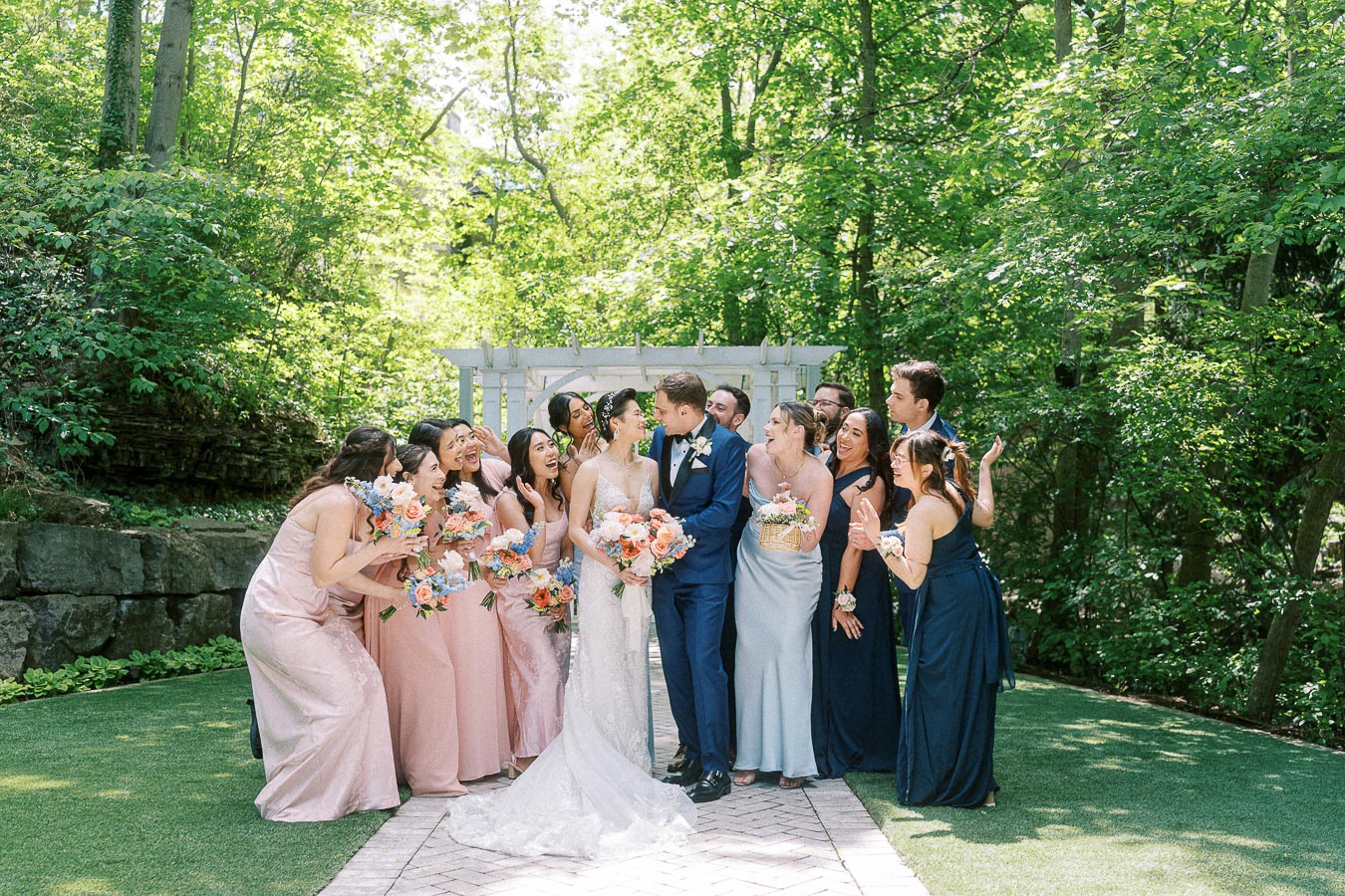 Miffy and Rawle kiss while their wedding party cheers under a white arbor in a wooded garden.