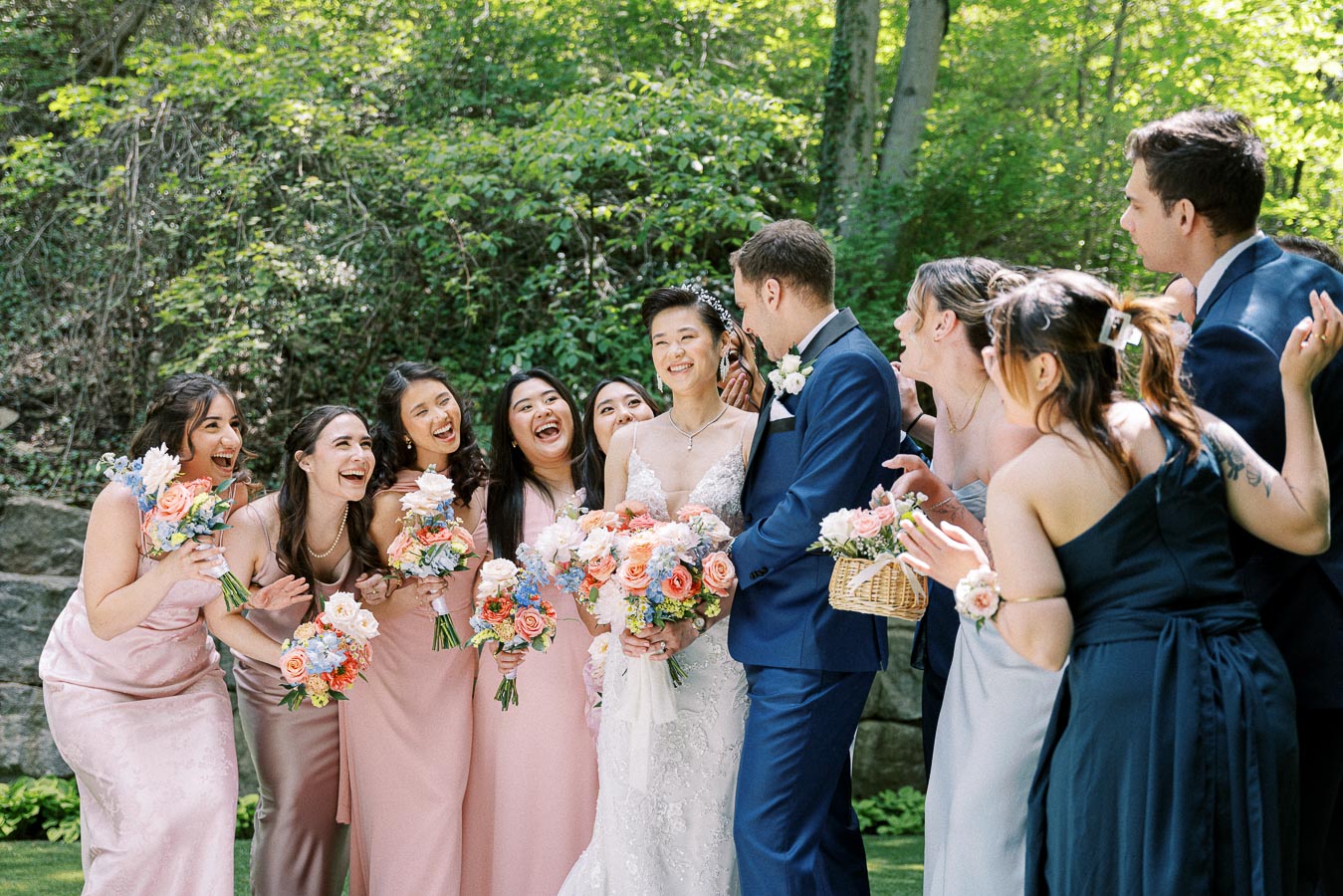 Bride and groom laugh with bridesmaids and groomsmen in a sunlit wooded setting at Ancaster Mill.