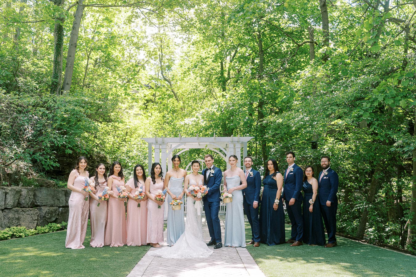 Wedding party standing with Miffy and Rawle under a white arbor in a leafy garden at Ancaster Mill