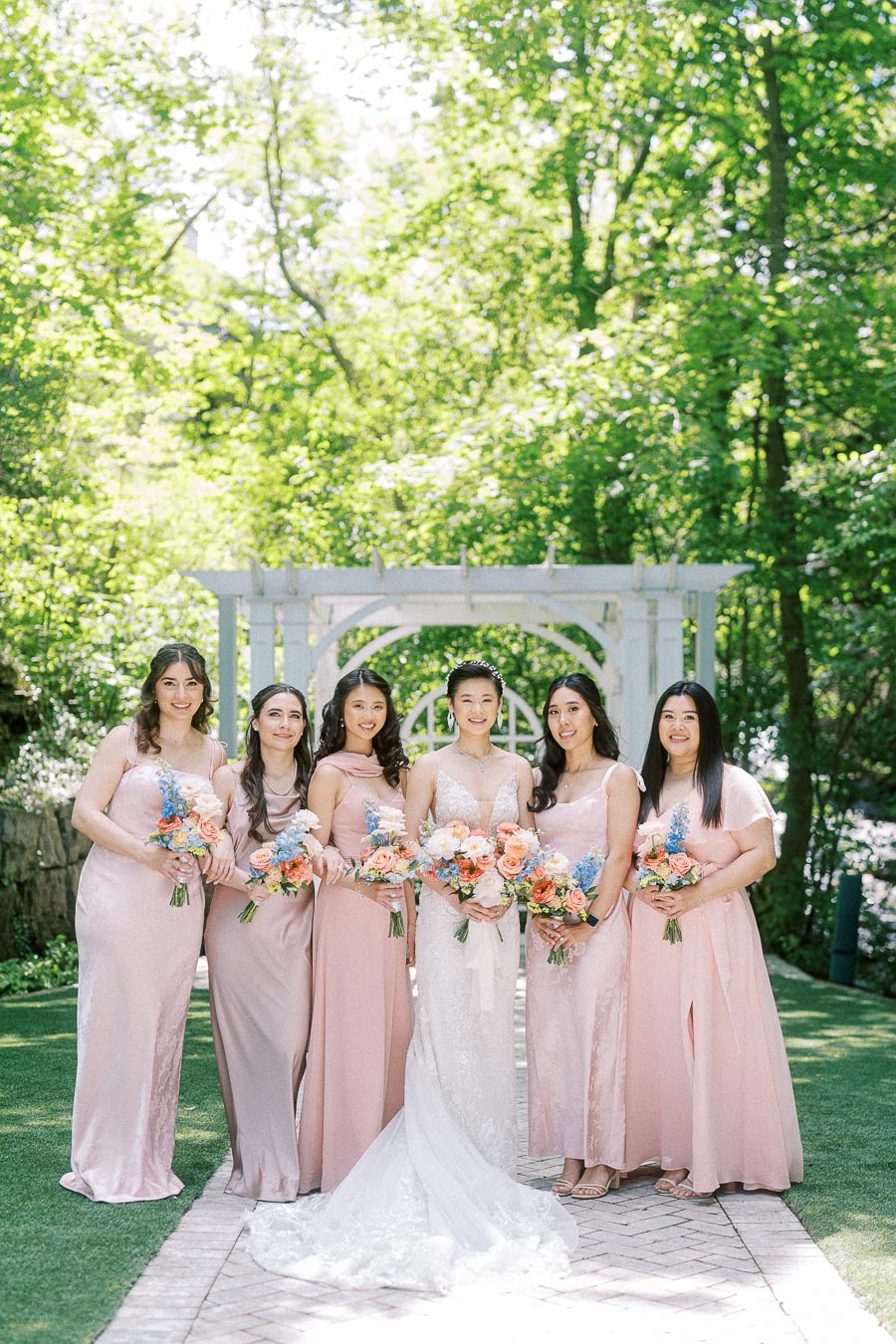 Bride Miffy stands with five bridesmaids in blush dresses holding bouquets beneath a white garden arbor.