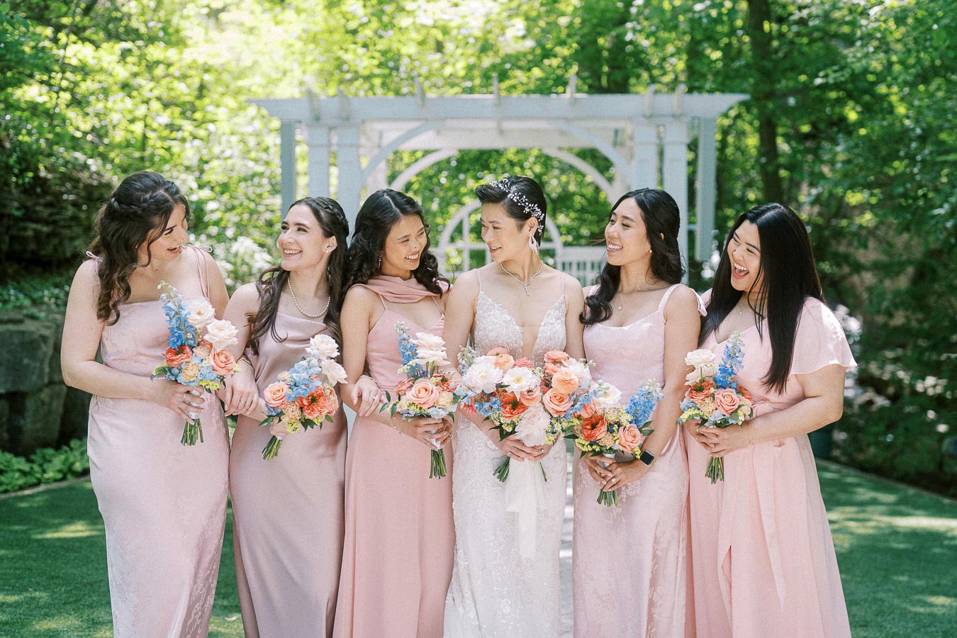 Bride in a lace gown laughs with five bridesmaids in blush dresses holding pastel bouquets under a white garden arbor.