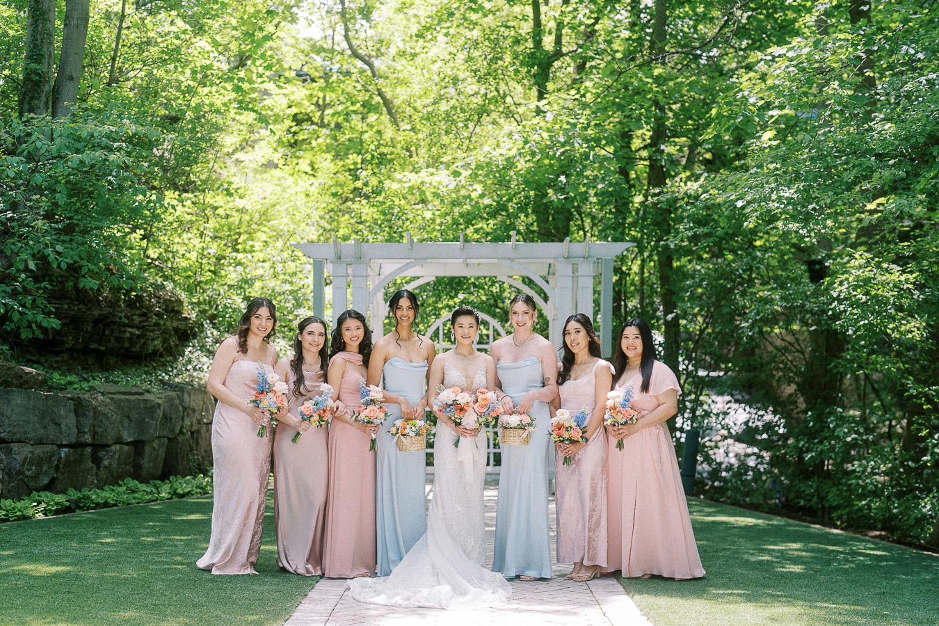 Miffy stands with seven bridesmaids in pastel dresses under a white arbor surrounded by green trees.