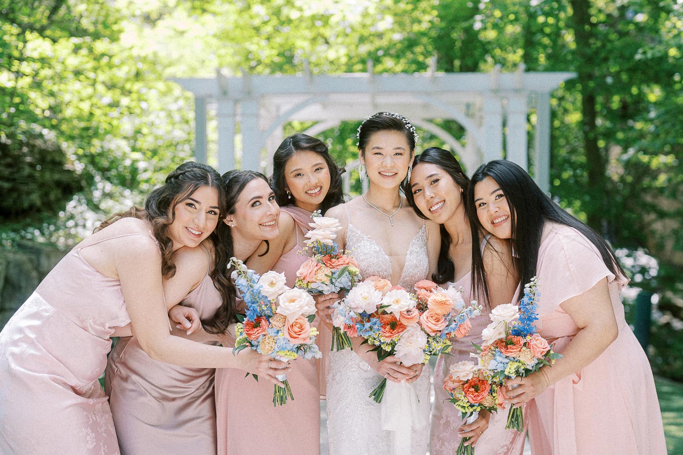 Miffy with five bridesmaids in blush pink dresses holding pastel bouquets under a white garden arbor.
