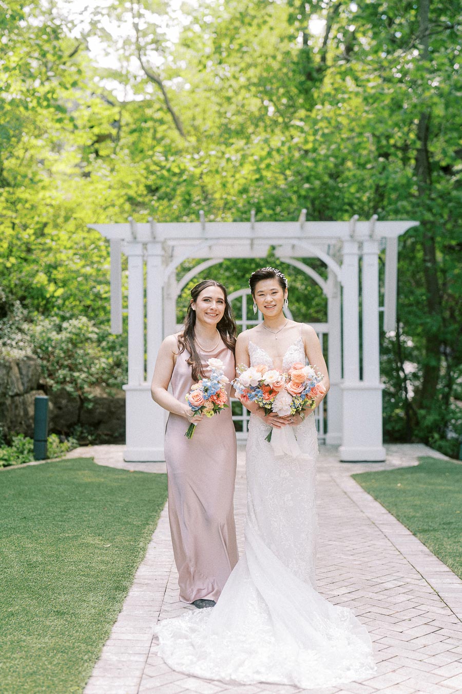 Bride in a lace gown and bridesmaid in blush walk with bouquets under a white garden pergola.