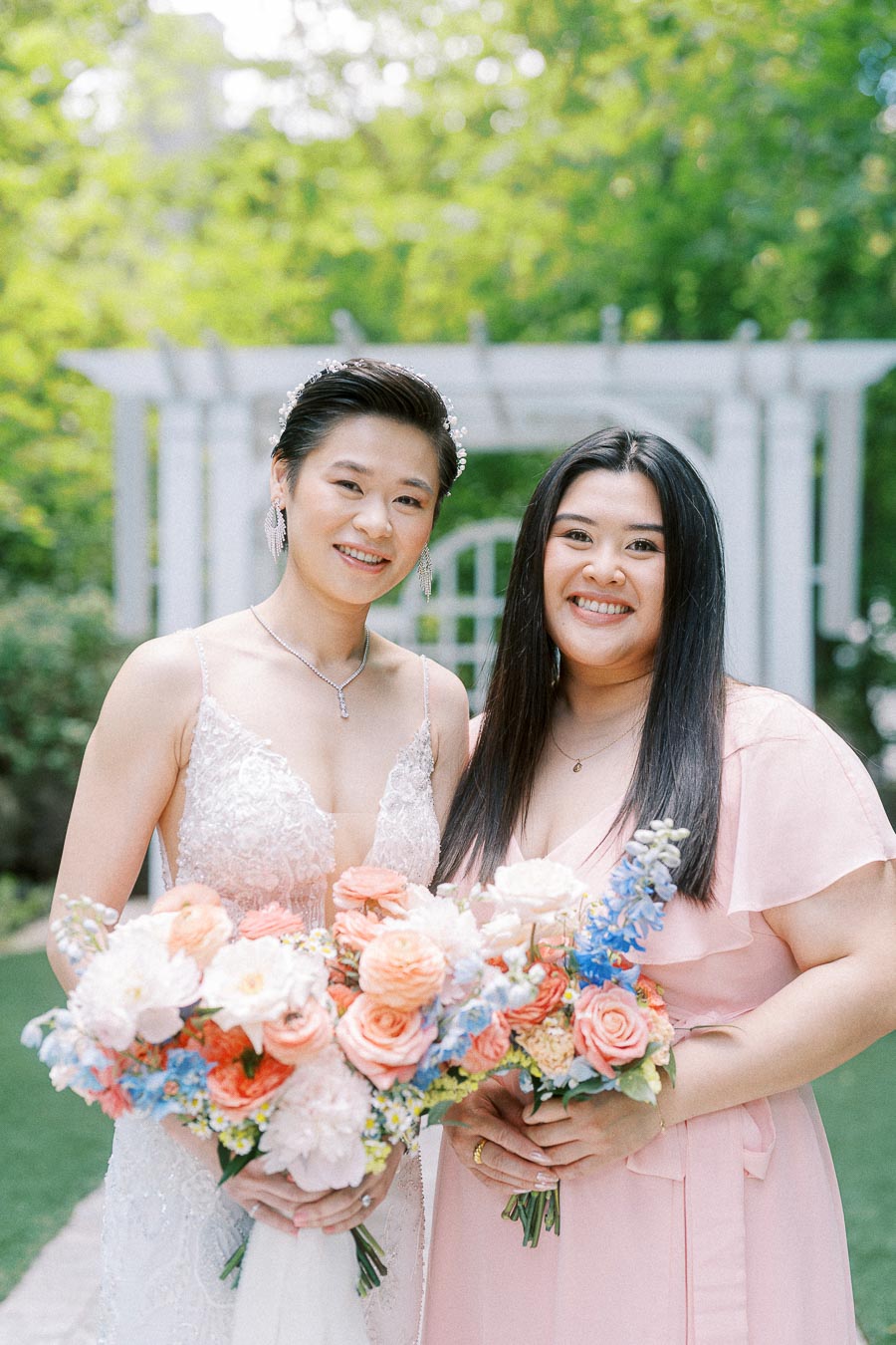 Miffy in a beaded wedding dress poses with a bridesmaid holding pastel bouquets in a garden.