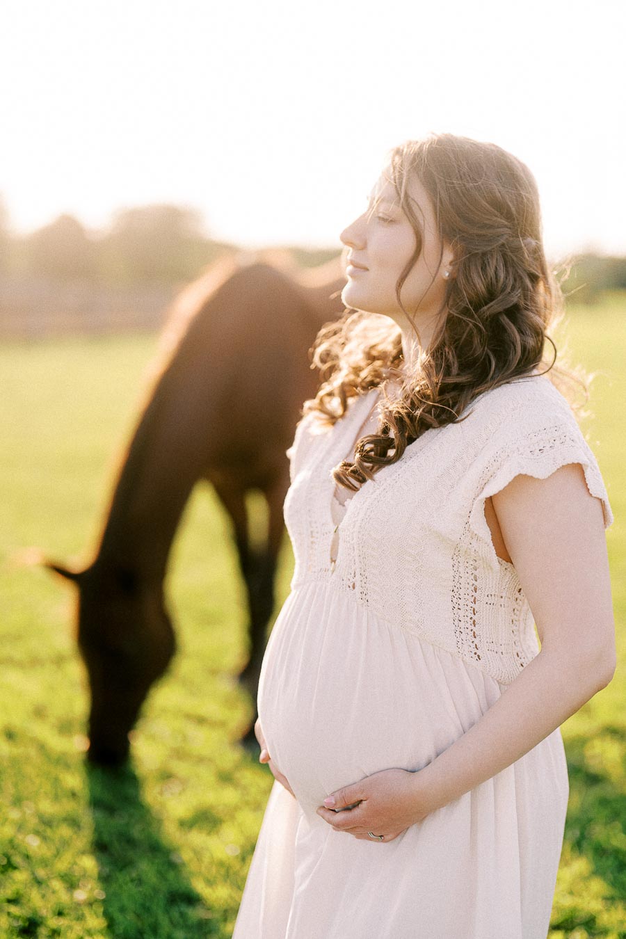 Pregnant woman in a cream dress with a grazing horse in a sunny field