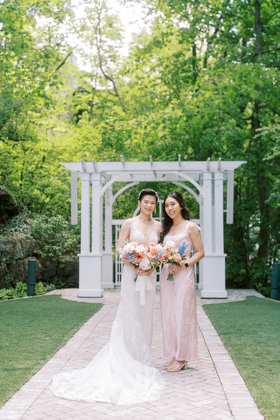Miffy in a lace wedding gown and a bridesmaid in pink holding bouquets beneath a white garden arbor.
