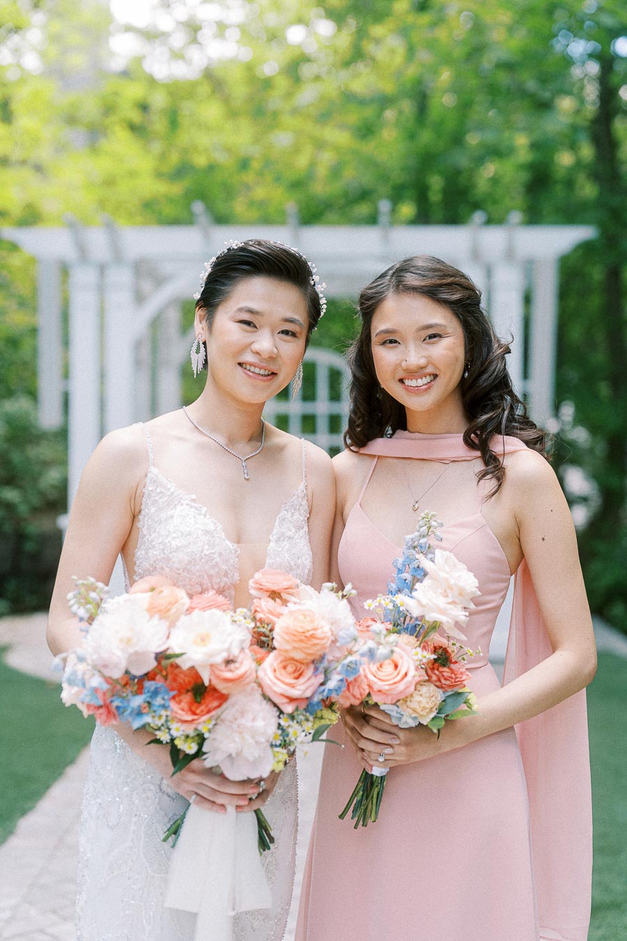 Miffy in a lace wedding dress and a bridesmaid in pink hold bouquets in a garden at Ancaster Mill.