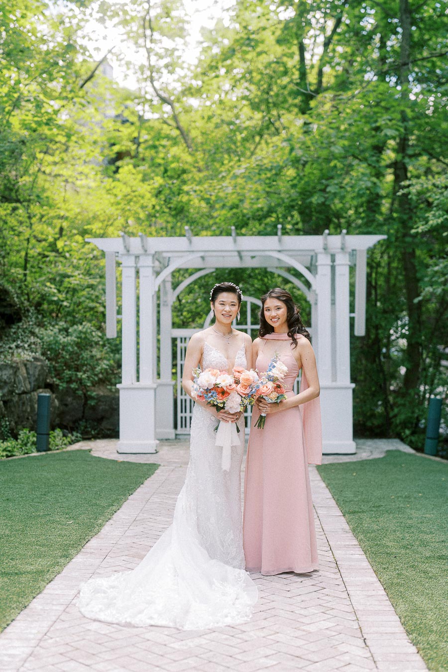 Bride Miffy and a bridesmaid in blush pink stand with bouquets before a white garden arbor at Ancaster Mill.
