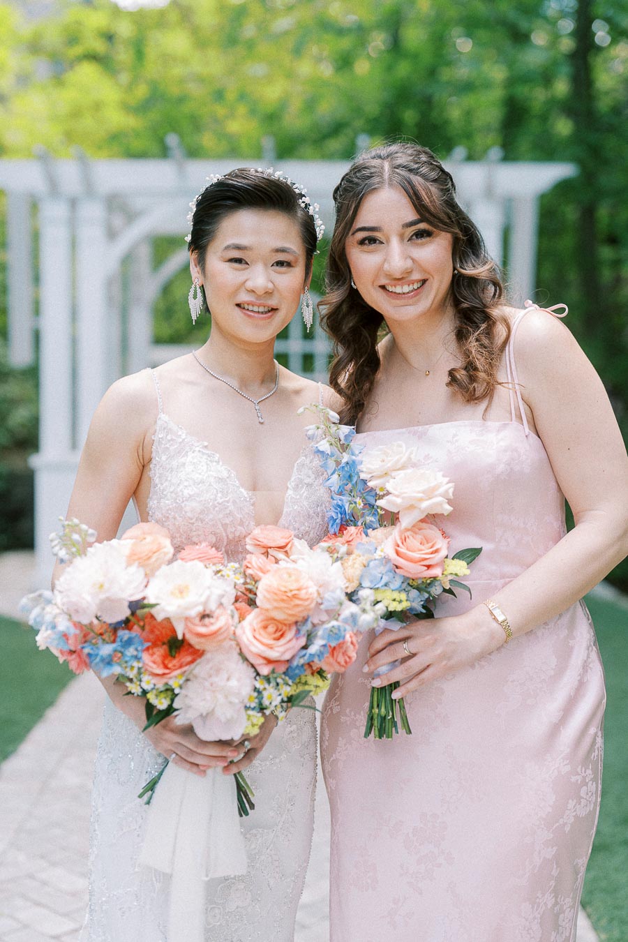 Bride Miffy and a bridesmaid hold pastel bouquets beside a white garden arbor at Ancaster Mill.