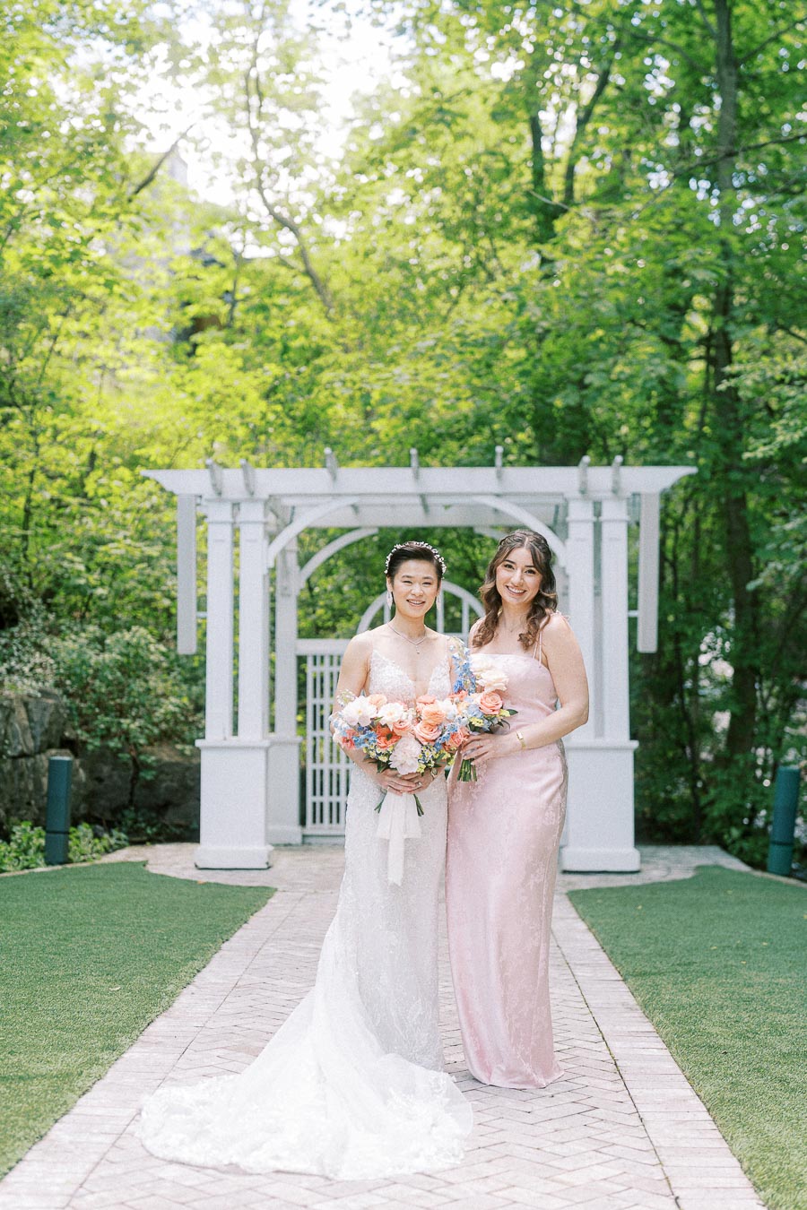 Bride and bridesmaid holding pastel bouquets on a garden walkway beneath a white pergola.