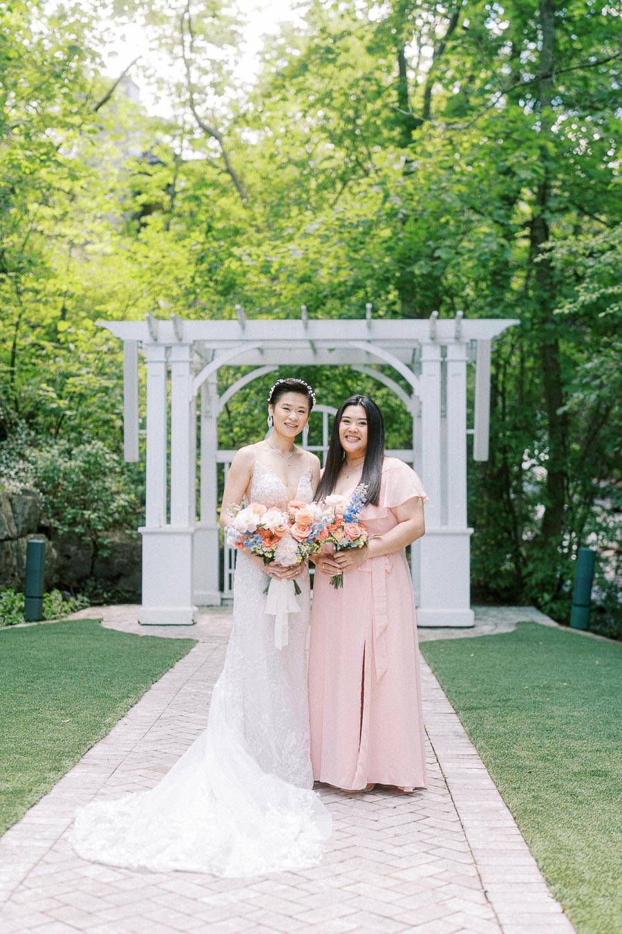Bride in a lace gown and woman in a pink dress hold bouquets before a white garden pergola.