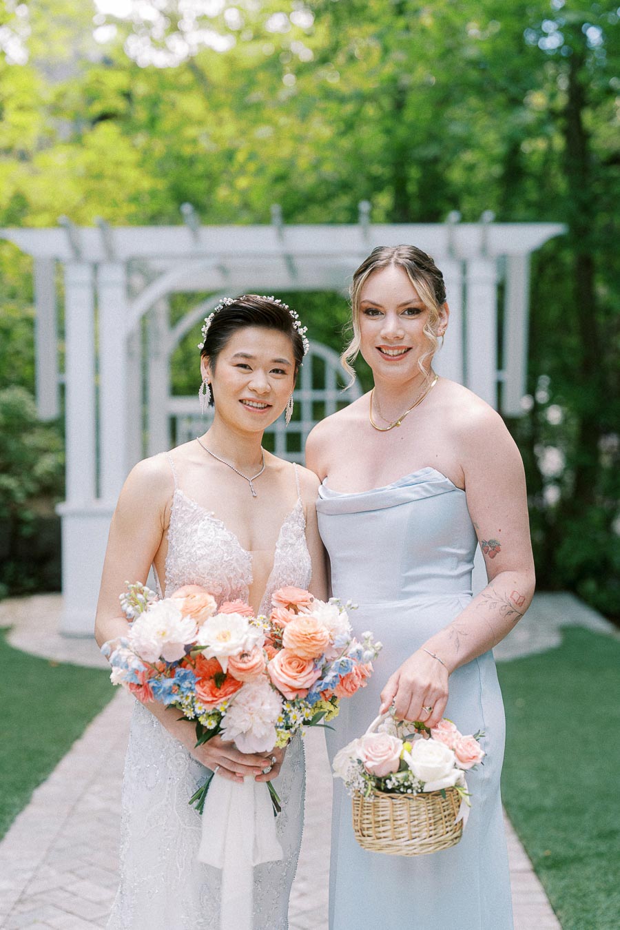 Miffy in a beaded wedding gown poses with a bridesmaid holding a flower basket in the Ancaster Mill garden.