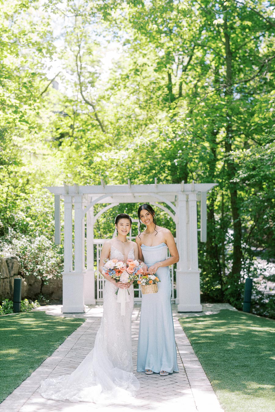 Bride and bridesmaid holding flowers beneath a white garden arbor at Ancaster Mill