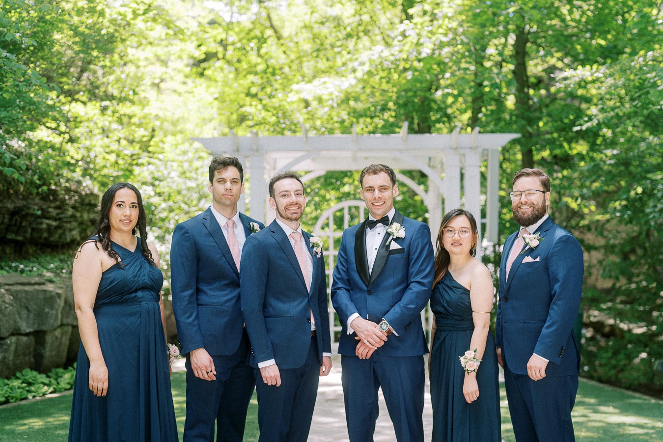 Rawle stands with five wedding party members in navy attire beneath a white garden pergola.
