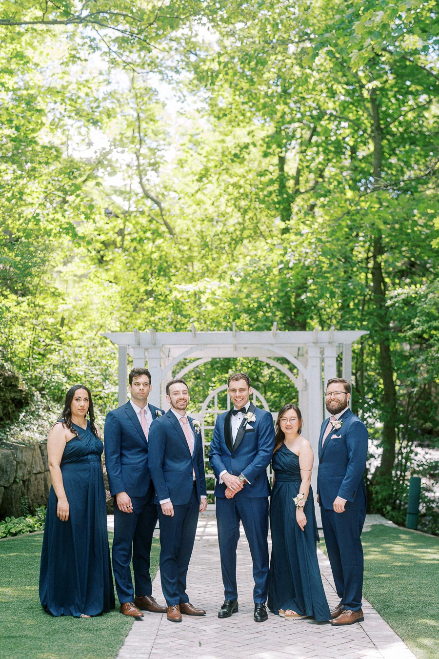 Wedding party in navy attire standing beneath a white arbor on a wooded path at Ancaster Mill