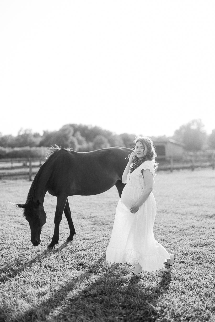 Pregnant woman in a white dress walking beside a grazing horse in a paddock